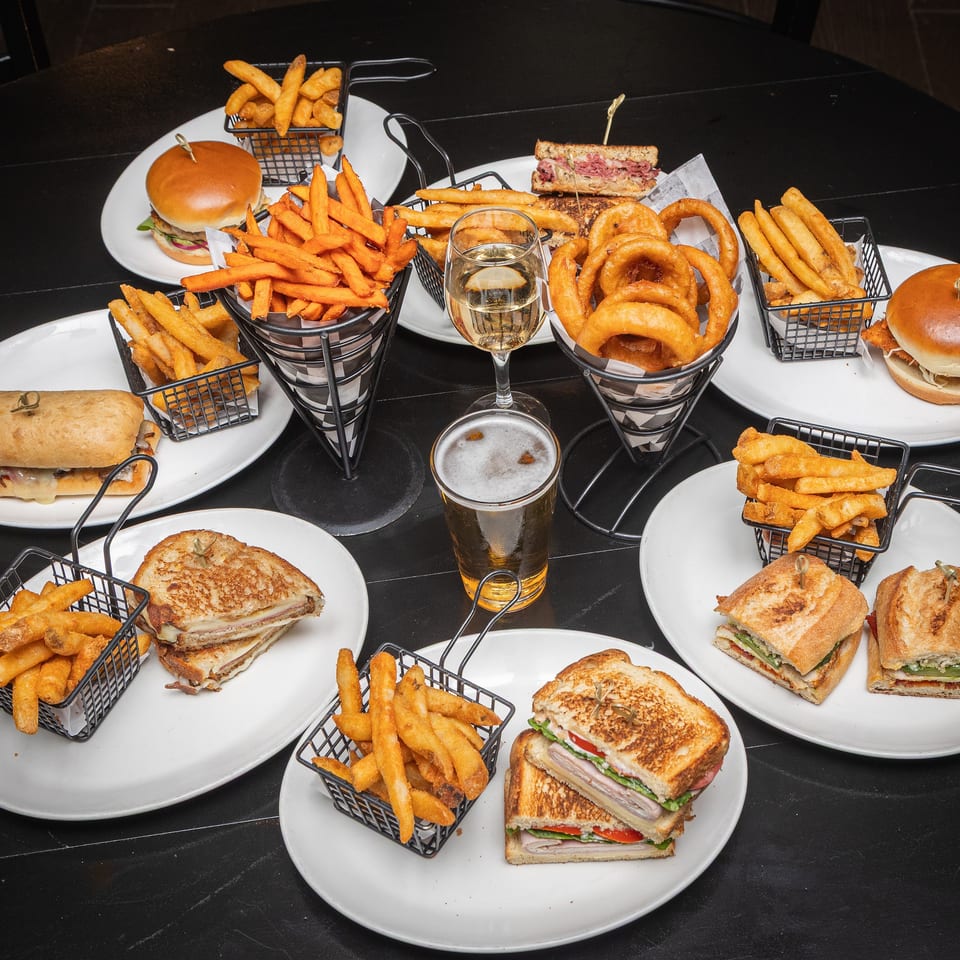 Assorted burgers, sandwiches, fries, onion rings, and drinks arranged on plates across a dark restaurant table.