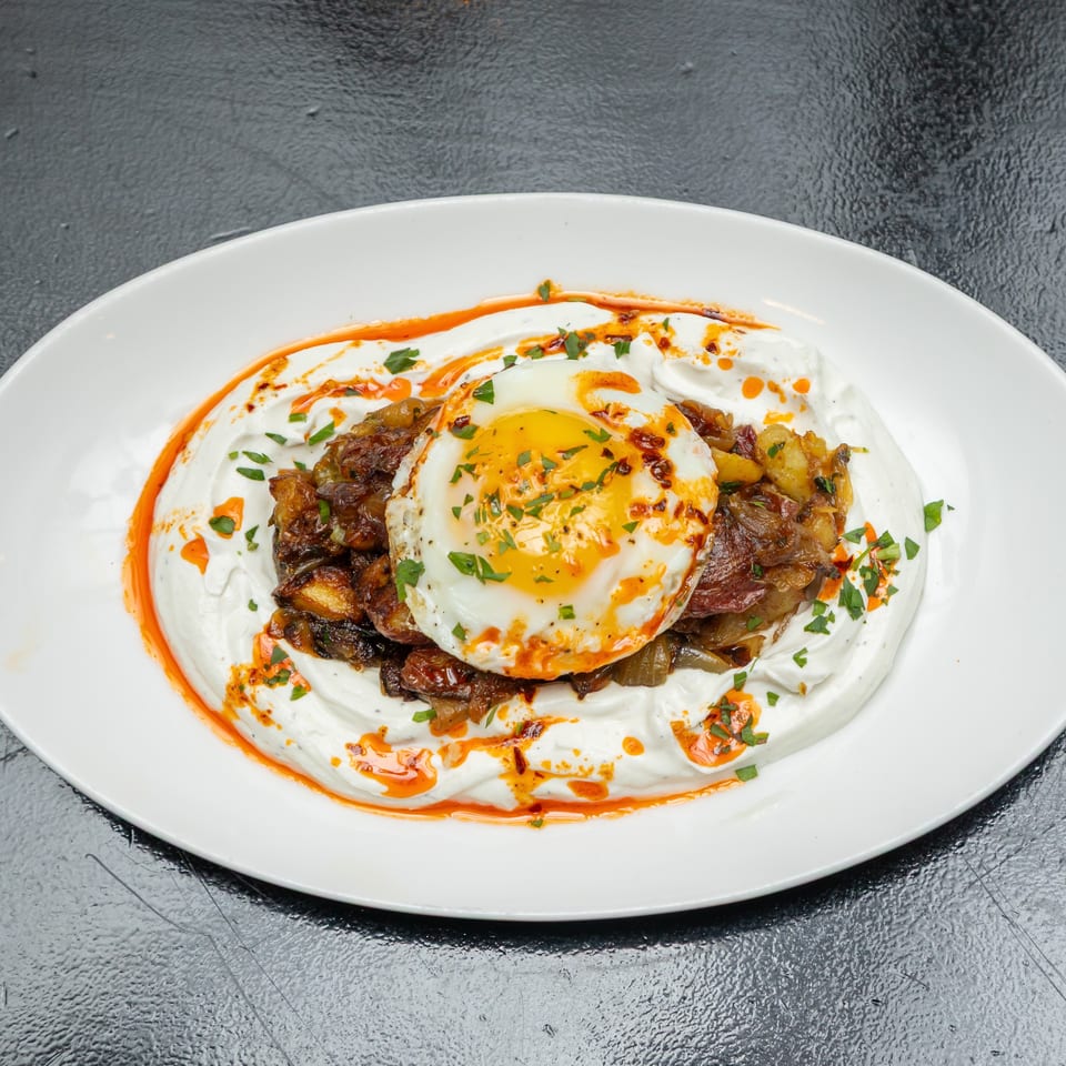Fried egg atop roasted potatoes and vegetables, served over creamy yogurt with herbs and chili oil on a white plate.