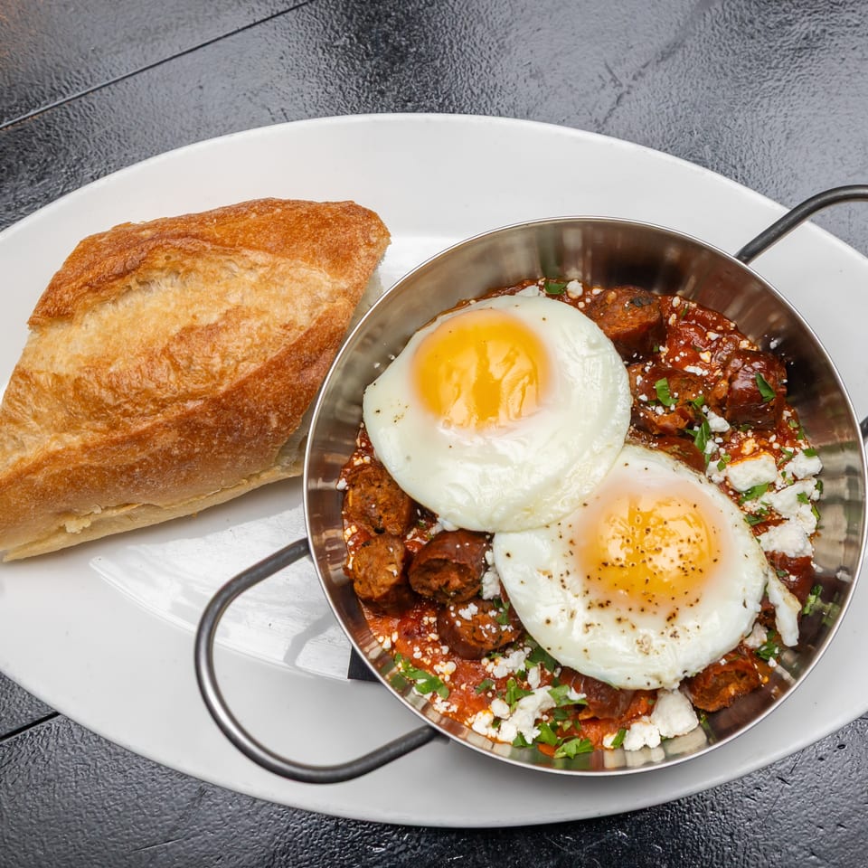 Skillet of meatballs in red sauce topped with two fried eggs, feta, and herbs, served with crusty bread.