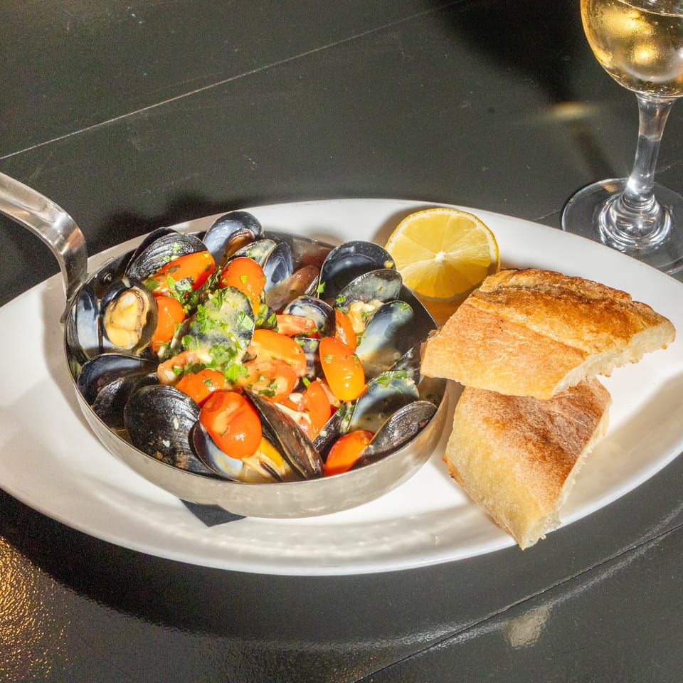 Bowl of mussels with tomatoes and herbs, toasted bread, lemon wedge, and a glass of white wine on a restaurant table.