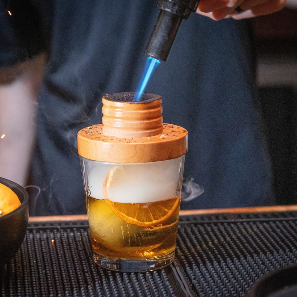 Bartender uses a blue flame to garnish an orange cocktail topped with smoke and a wooden coaster.