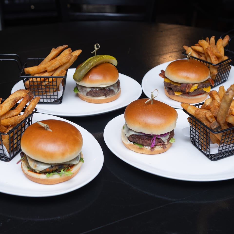 Four gourmet burgers with varied toppings and baskets of golden fries arranged on white plates over a dark table.