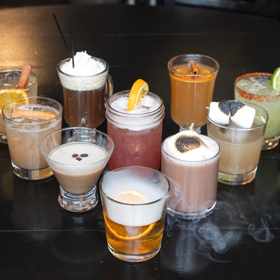 Assortment of colorful cocktails garnished with citrus, cinnamon, whipped cream, marshmallows, and toasted toppings on a dark bar.