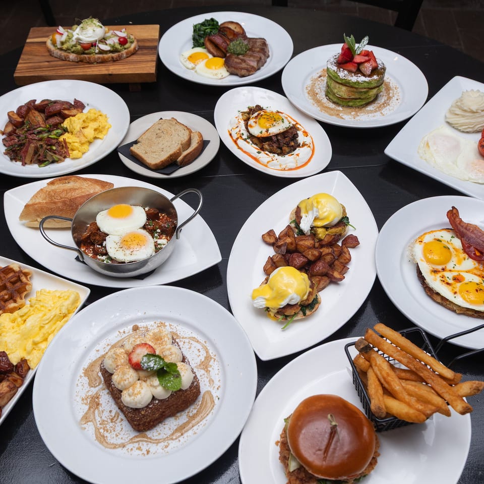 Overhead spread of restaurant brunch dishes including eggs, bacon, steak, pancakes, waffles, avocado toast, burgers, and fries.