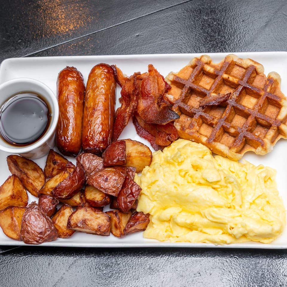 Breakfast platter with waffles, sausages, bacon, scrambled eggs, roasted potatoes, and syrup on a white rectangular plate.