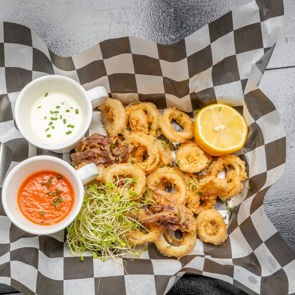Crispy fried calamari served with lemon, microgreens, creamy herb dip, and spicy red sauce on checkered paper.