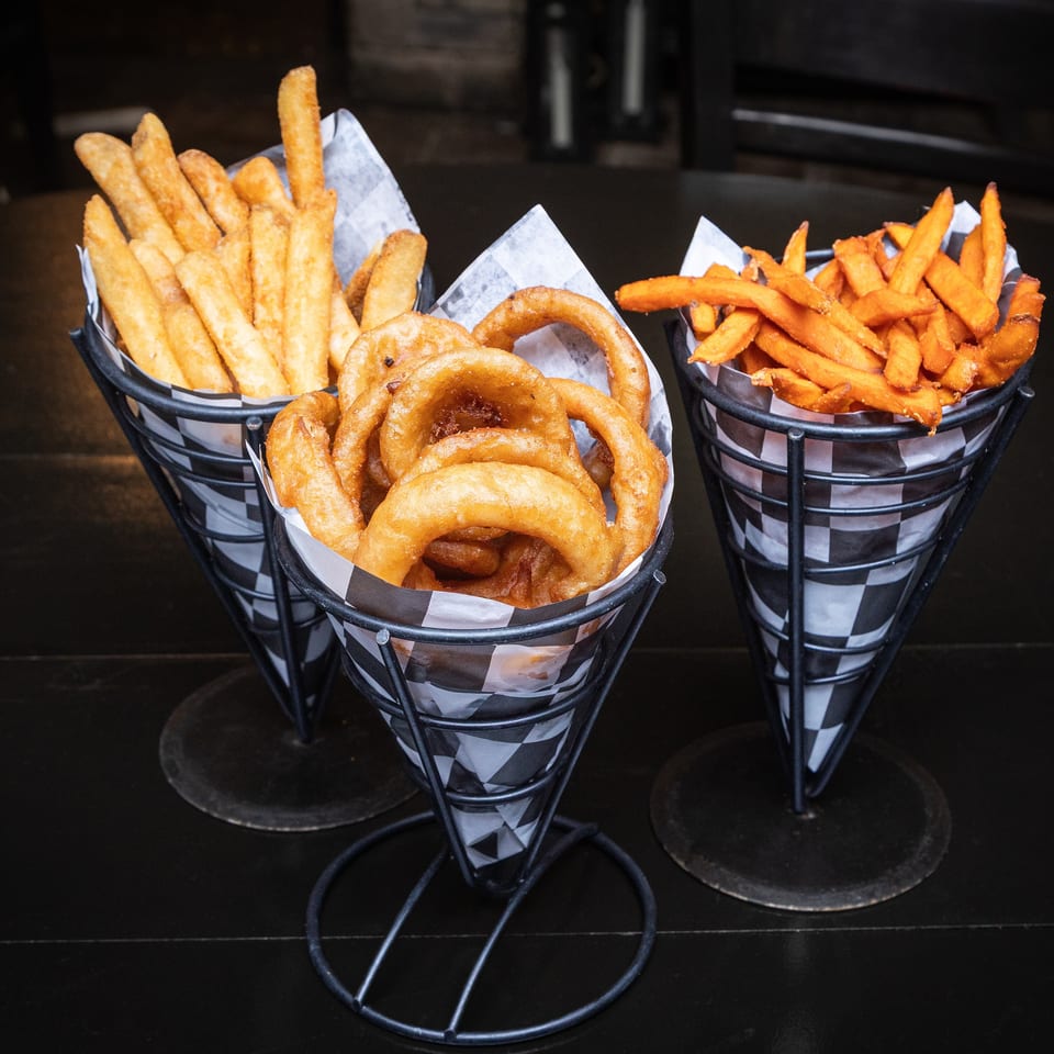 Three checkered baskets hold regular fries, onion rings, and sweet potato fries on a dark restaurant table.