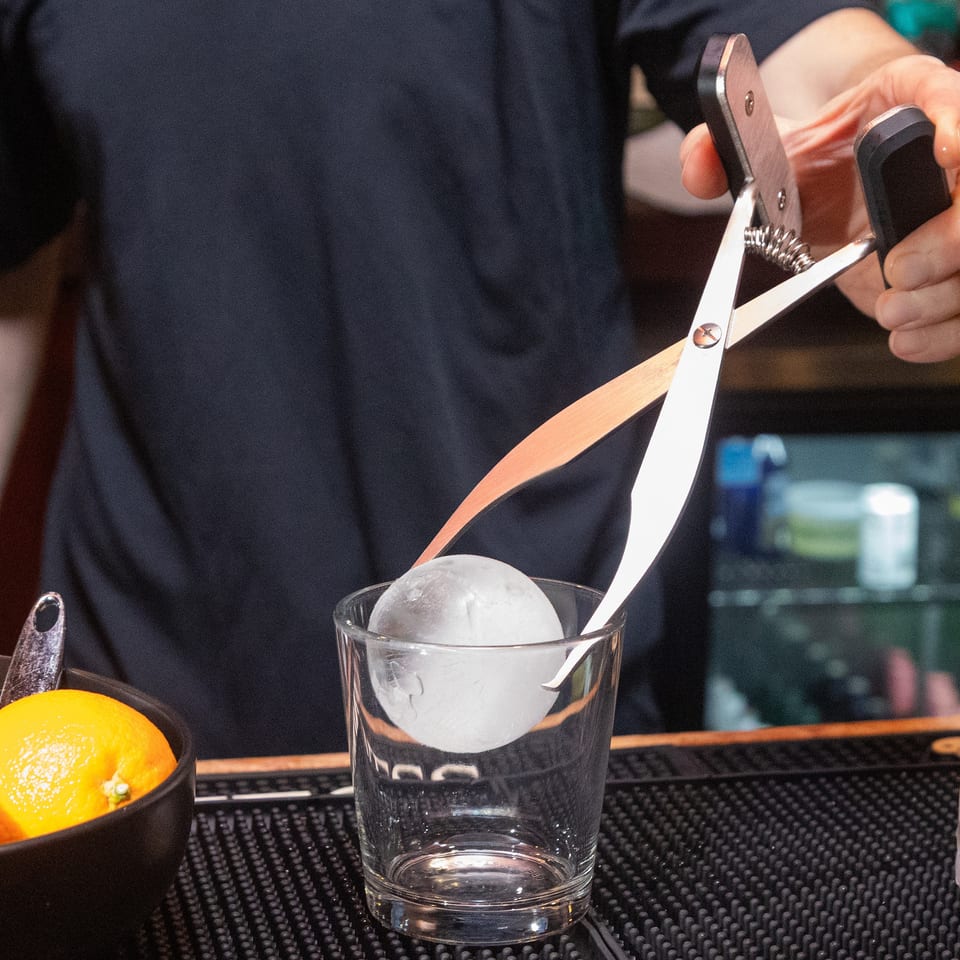 Bartender uses large scissors to cut an orange peel over a glass containing a large ice sphere.