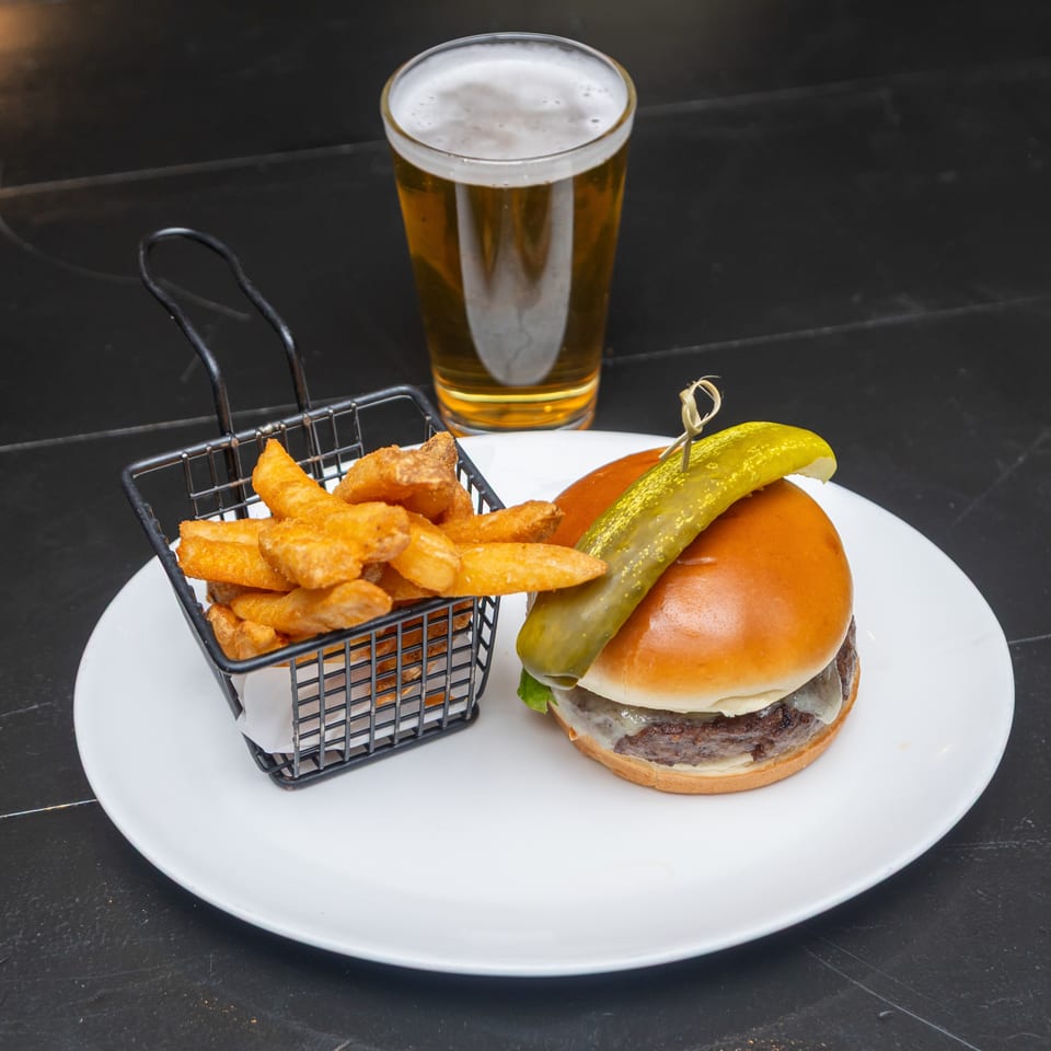 Cheeseburger with a pickle spear and fries in a wire basket, served on a white plate with a foamy beer.