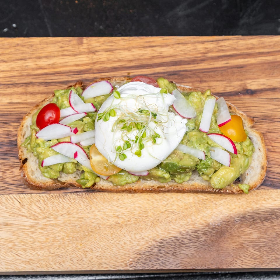 Open-faced toast topped with mashed avocado, radishes, cherry tomatoes, burrata, and microgreens on a wooden board.