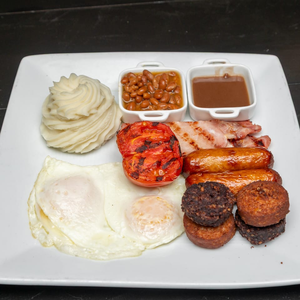 Hearty breakfast platter with mashed potatoes, fried eggs, grilled tomato, bacon, sausages, beans, and brown sauce.