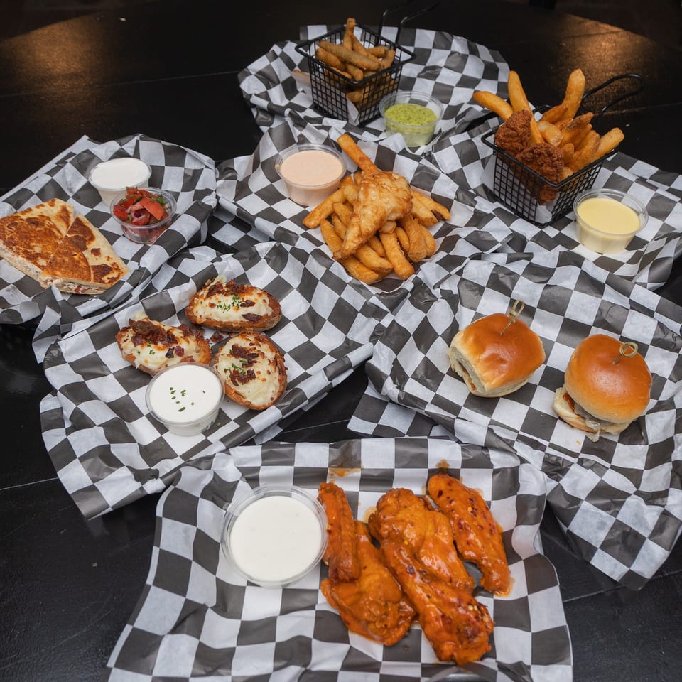 A restaurant spread features fries, fried chicken, sliders, quesadillas, cheesy appetizers, sauces, and buffalo wings on checkered paper.