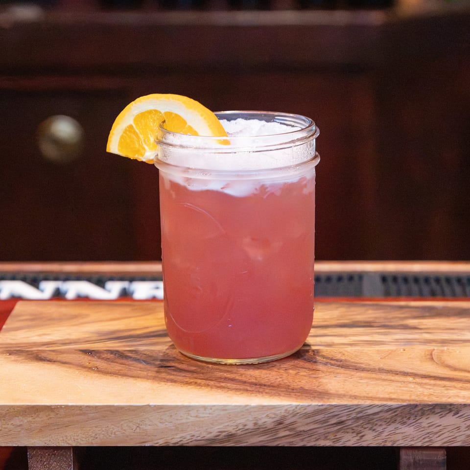 Pink iced cocktail garnished with an orange slice, served in a glass on a wooden bar.