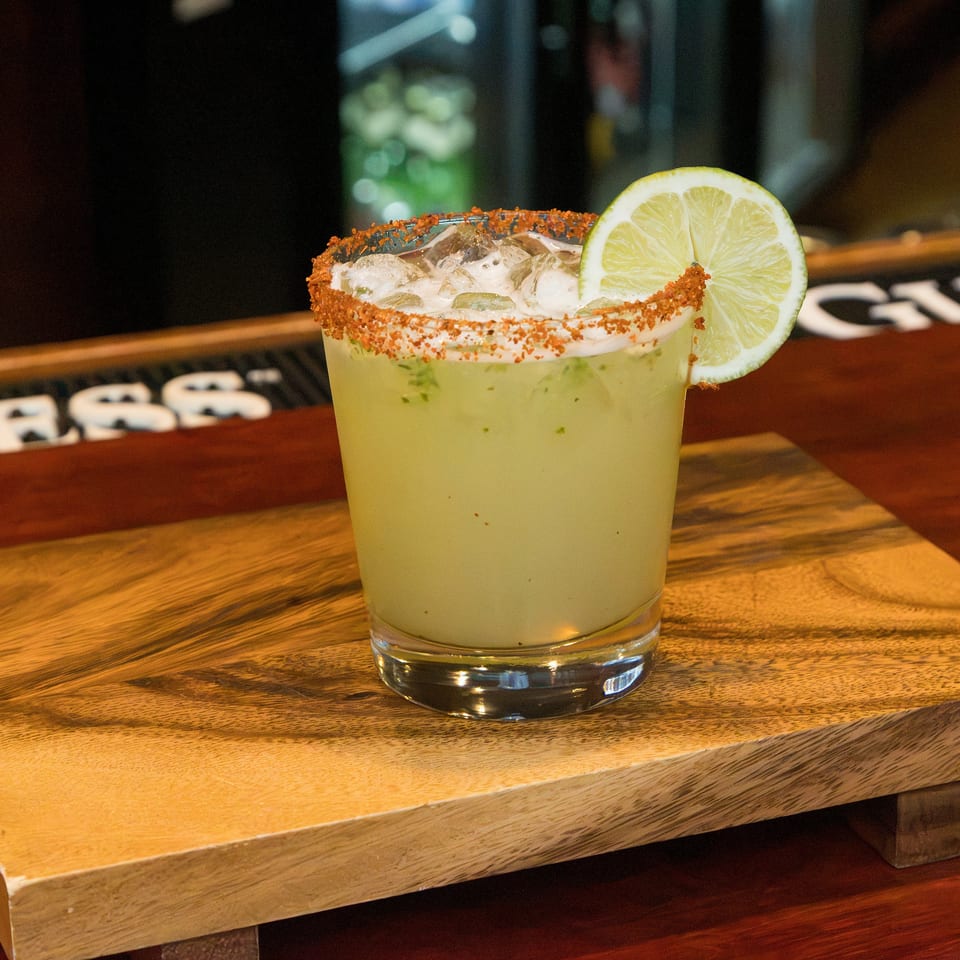 Pale green cocktail with ice, a chili-salt rim, and lime garnish sits on a wooden board at a dimly lit bar.
