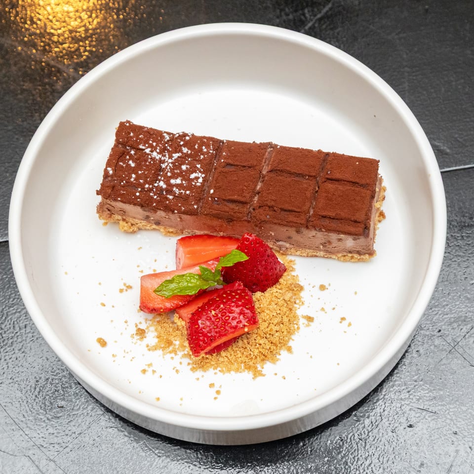 Chocolate dessert bar dusted with cocoa and powdered sugar, served with strawberries, mint, and biscuit crumbs on a white plate.