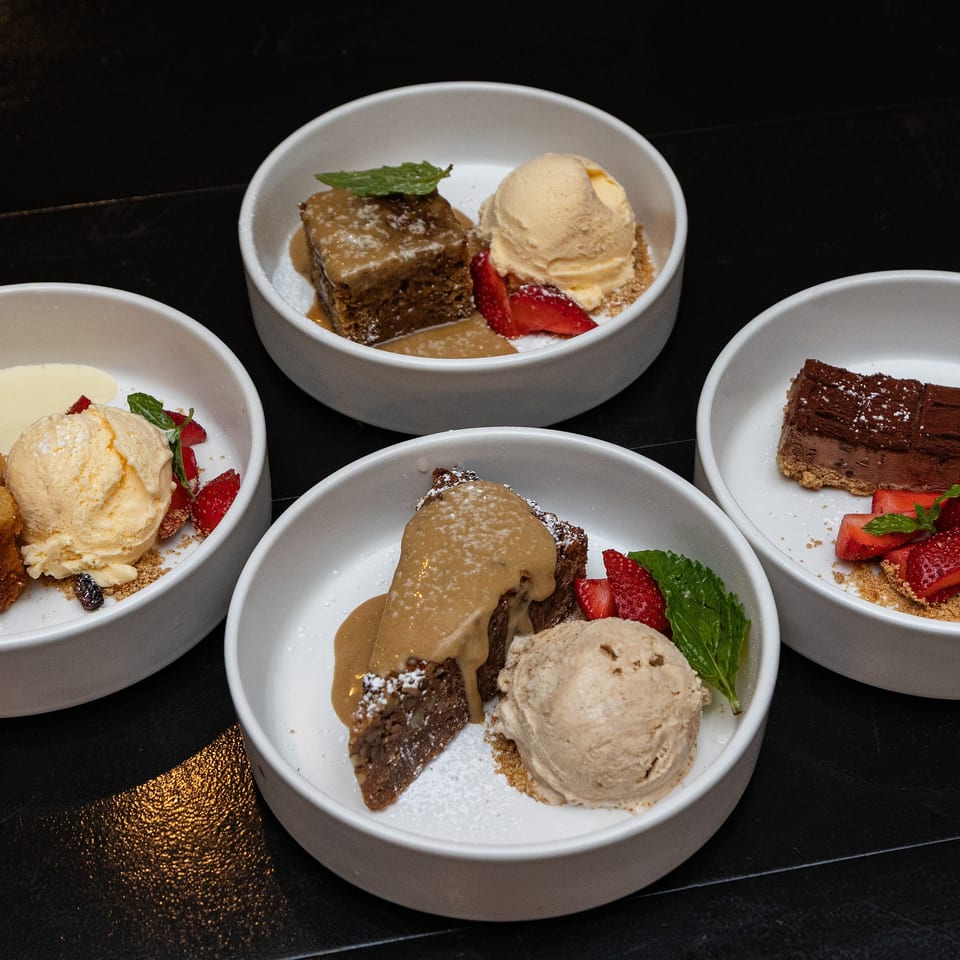 Four white bowls of assorted desserts, including cakes, ice cream, strawberries, mint, and powdered sugar, arranged on a black table.
