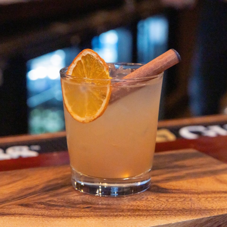 Citrus cocktail in a low glass, garnished with a dried orange slice and cinnamon stick on a wooden bar.