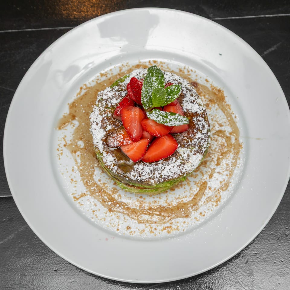 Stacked dessert topped with sliced strawberries, fresh mint, powdered sugar, and syrup on a white plate.