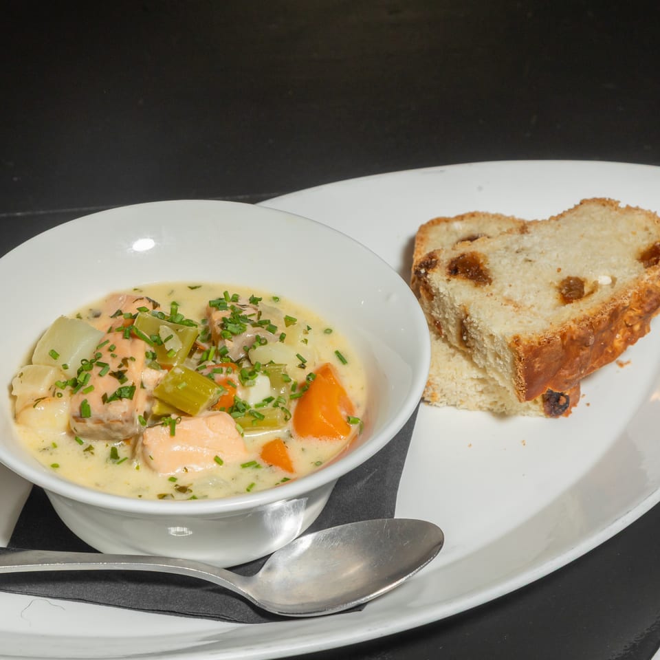 Creamy salmon and vegetable soup garnished with chives, served with raisin bread slices on a white plate.