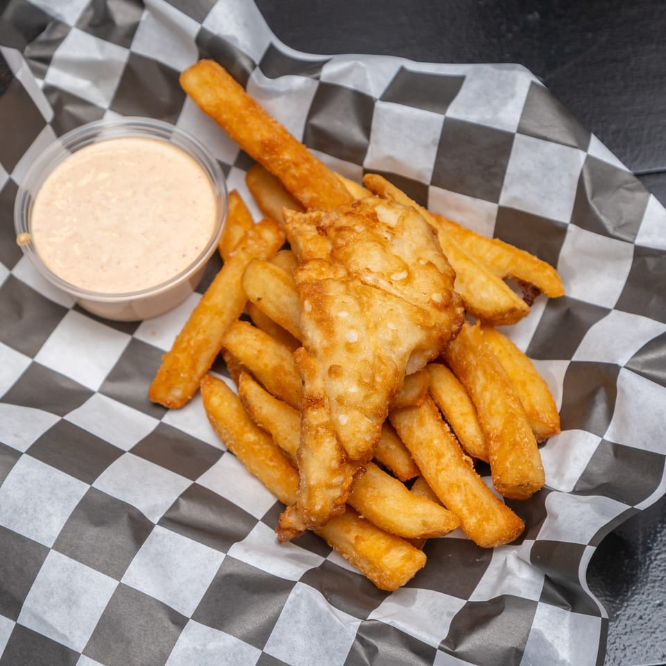 Golden fried fish and thick-cut fries served with creamy dipping sauce on black-and-white checkered paper.