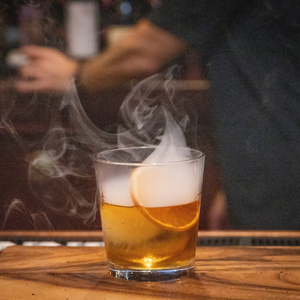 Whiskey cocktail with an orange slice and foamy top, steaming in a glass on a wooden restaurant table.