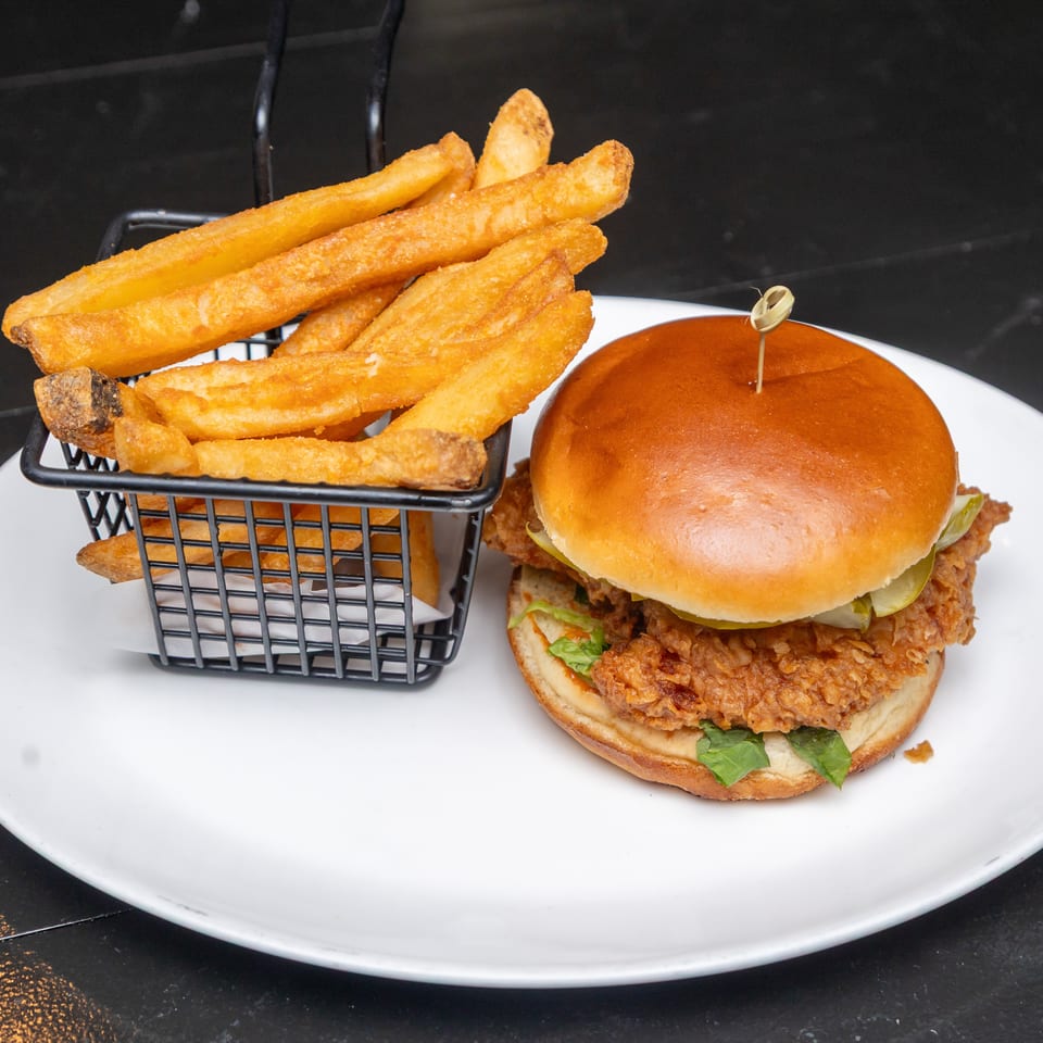 Crispy fried chicken sandwich with lettuce on a brioche bun, served alongside golden fries in a basket on a white plate.