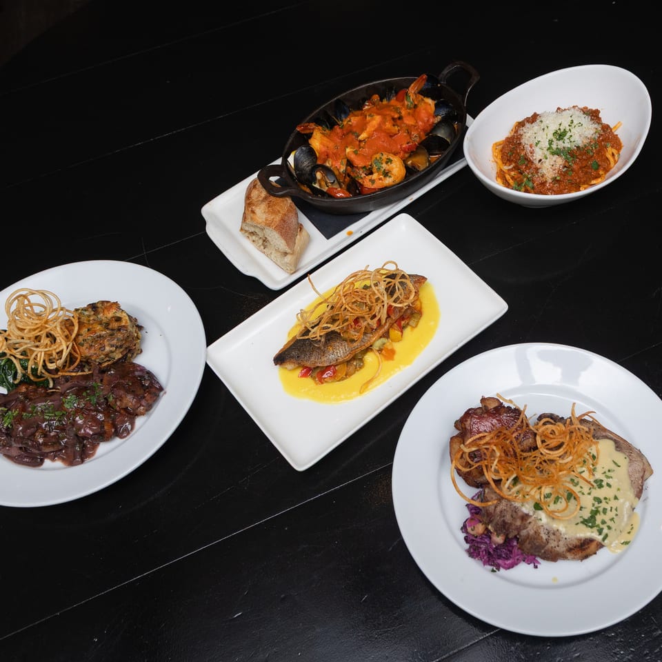 Assortment of plated restaurant dishes, including seafood, pasta, steak, fish, crispy noodles, vegetables, and bread on a black table.